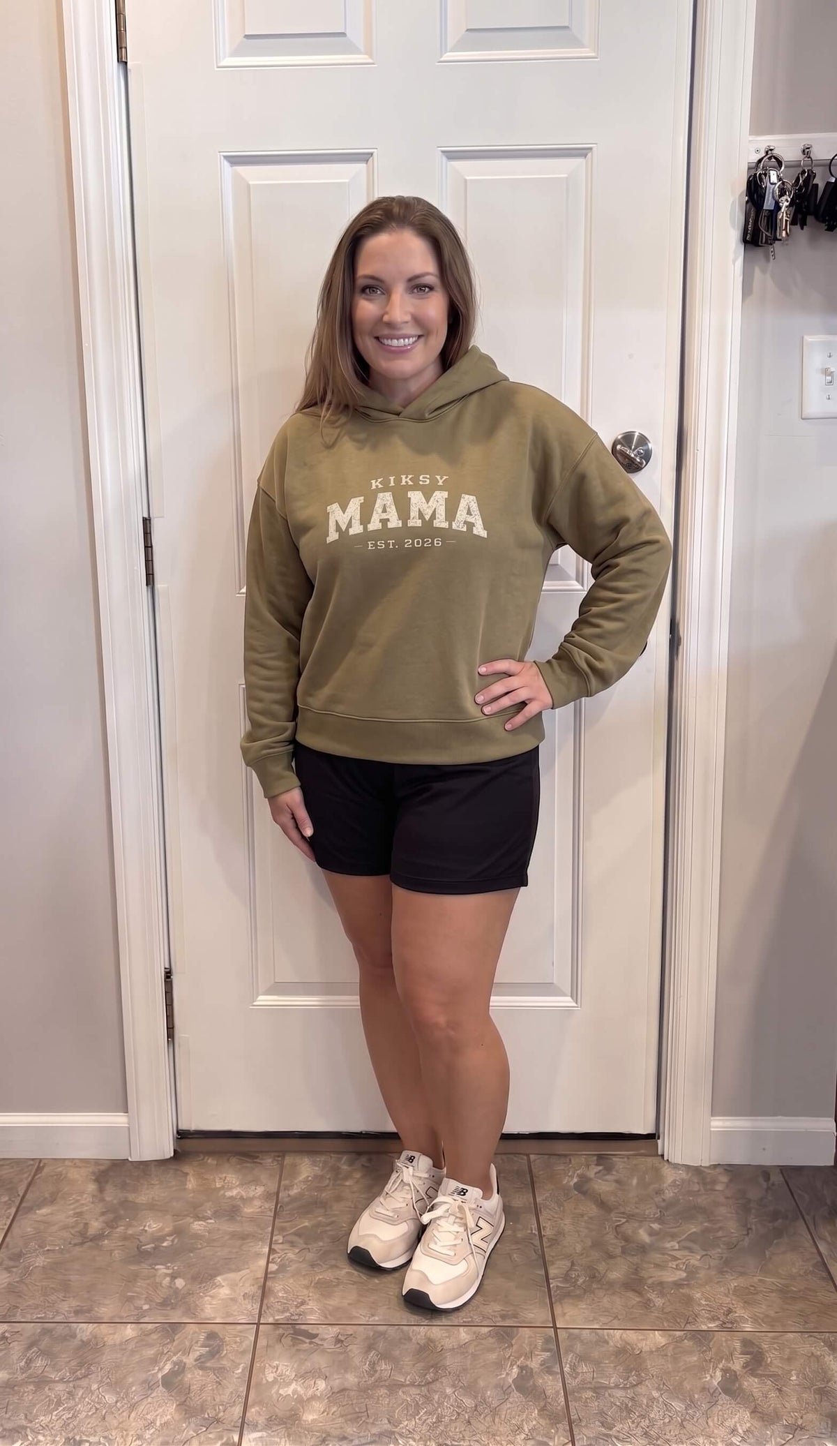 Woman wearing a Kiksy Mama Est 2026 hoodie with distressed white varsity lettering.