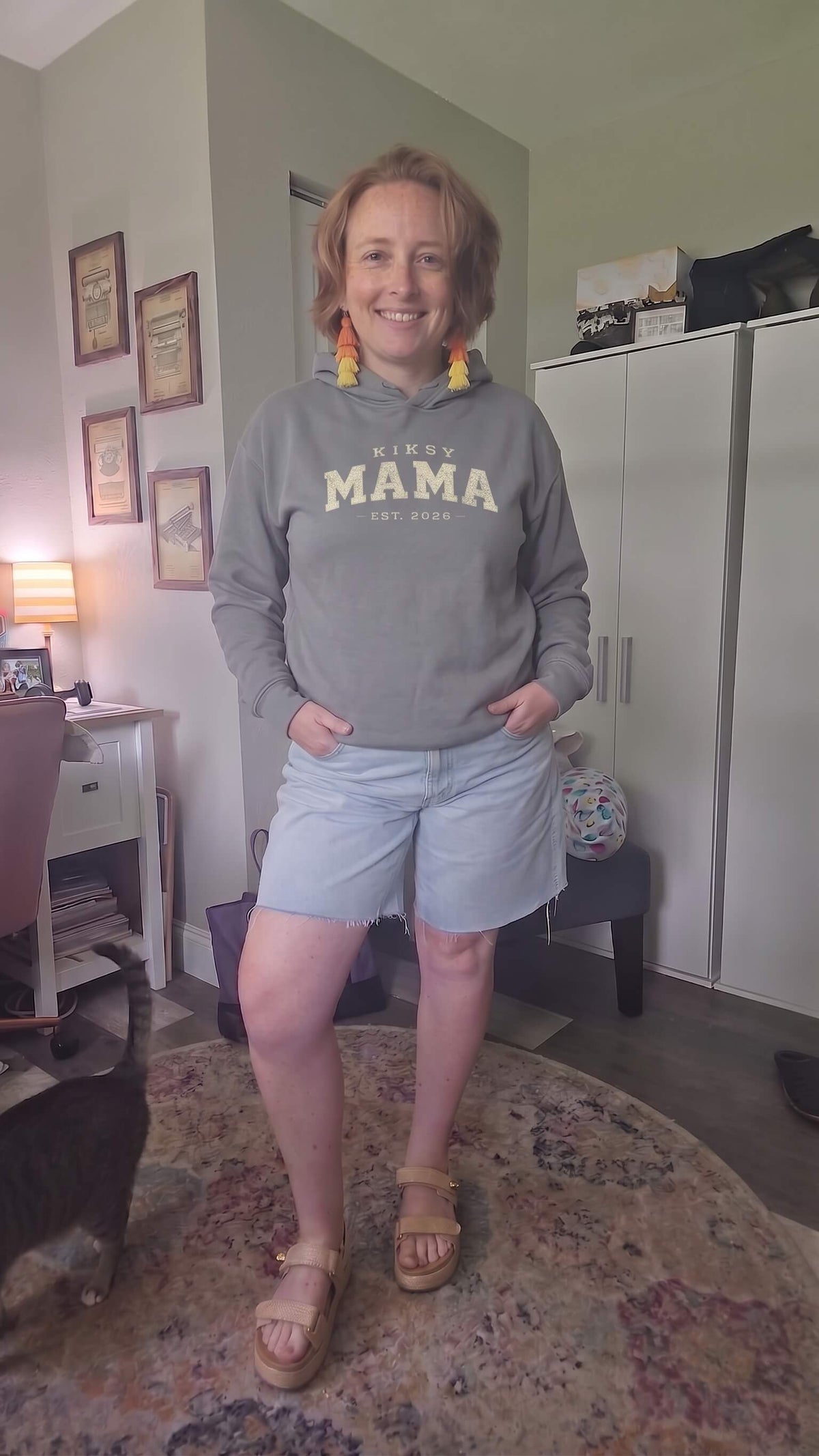 Woman wearing a Kiksy Mama Est 2026 hoodie with distressed white varsity lettering.