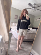 Woman wearing a black Kiksy Mama Est 2026 hoodie with distressed white varsity lettering.