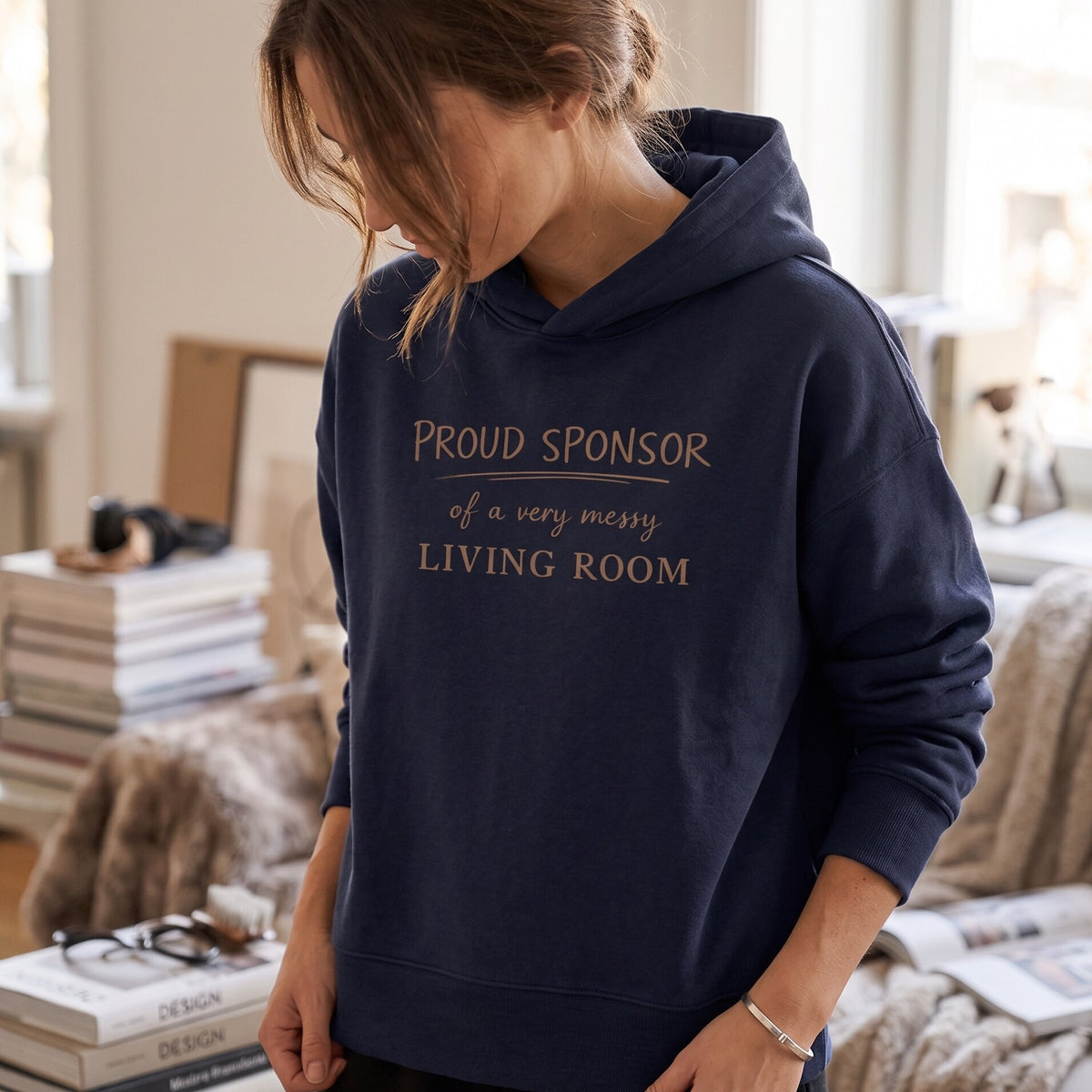 Woman wearing Kiksy dark blue hoodie featuring the text Proud Sponsor of a very messy LIVING ROOM