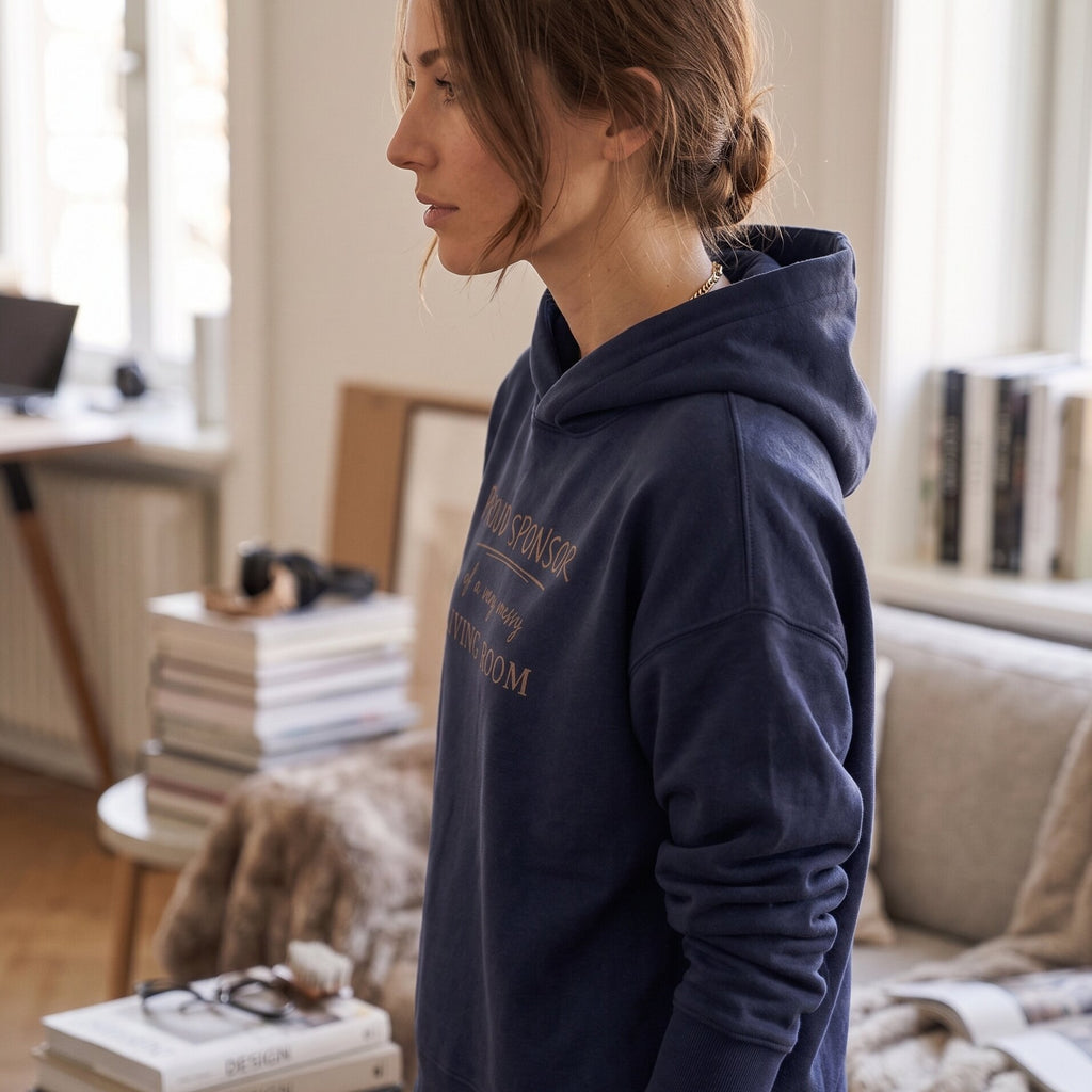 Woman wearing Kiksy dark blue hoodie featuring the text Proud Sponsor of a very messy LIVING ROOM