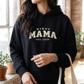 Kiksy black Mama Est 2026 hoodie worn by a woman indoors, featuring vintage varsity lettering and cozy fleece fabric.