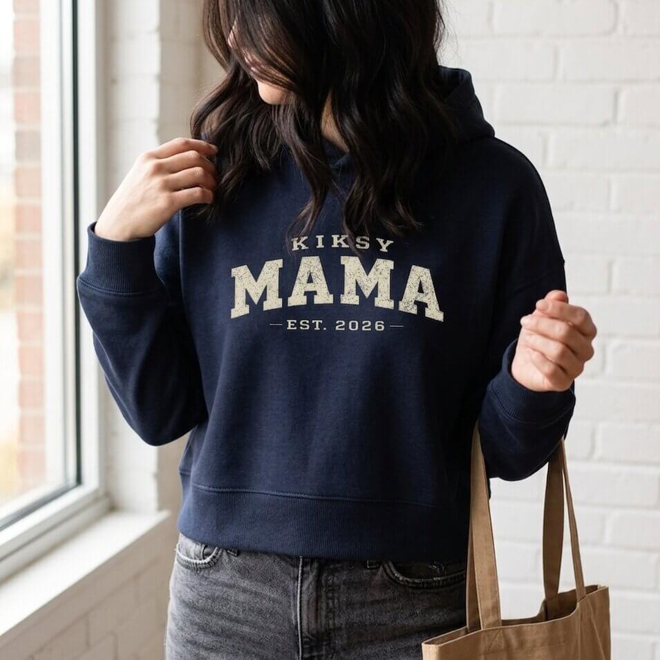 Kiksy navy Mama Est 2026 hoodie worn by a woman indoors, featuring vintage varsity lettering and cozy fleece fabric.
