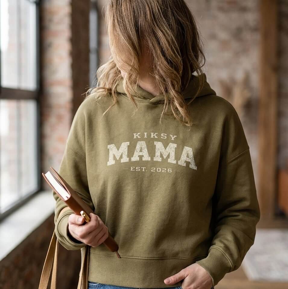 Kiksy olive Mama Est 2026 hoodie worn by a woman indoors, featuring vintage varsity lettering and cozy fleece fabric.