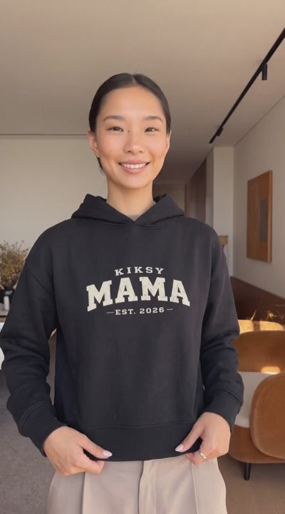 Woman wearing a black Kiksy Mama Est 2026 hoodie with distressed white varsity lettering.